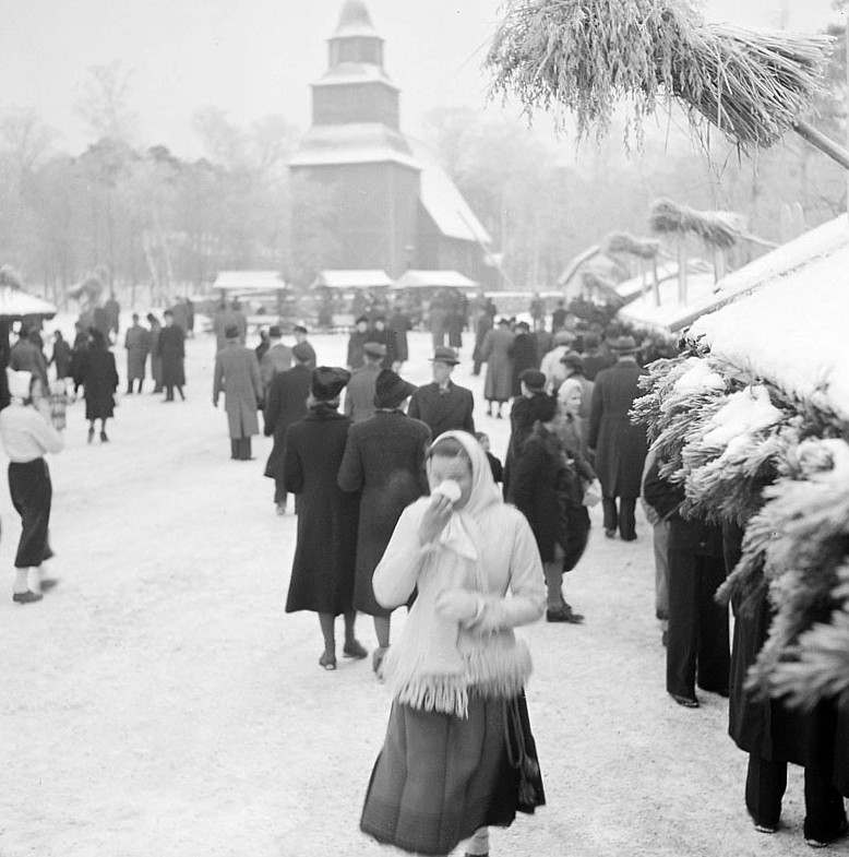 Black-and-white photograph of Skansen's Christmas market in the 1940s with stalls, bonfire, and visitors in winter coats